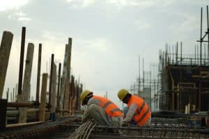 Two construction workers in hard hats and safety vests tying rebar on a building site
