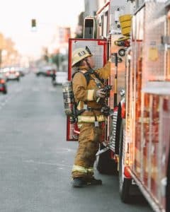 A firefighter in full turnout gear and air pack stepping off the back of an engine